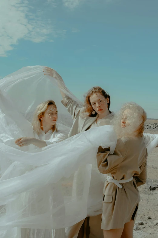 a group of women standing on top of a sandy beach, an album cover, by Sara Saftleven, pexels contest winner, surrealism, white cloth in wind shining, three women, robe. perfect faces, concert