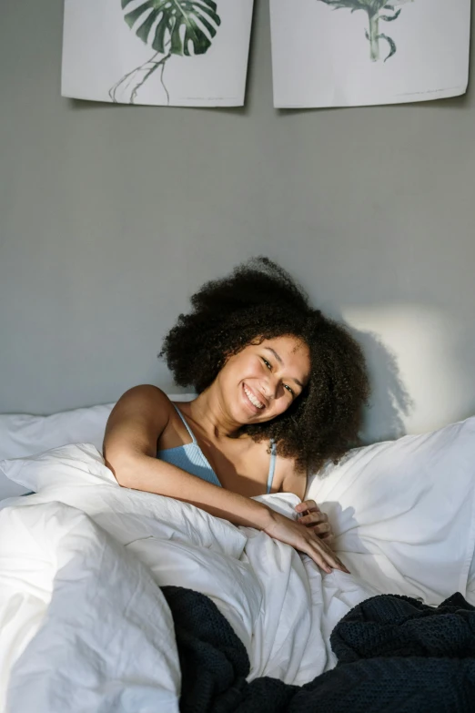 a woman laying in bed with a white comforter, a cartoon, trending on pexels, dark short curly hair smiling, black young woman, bright sunshine, angled