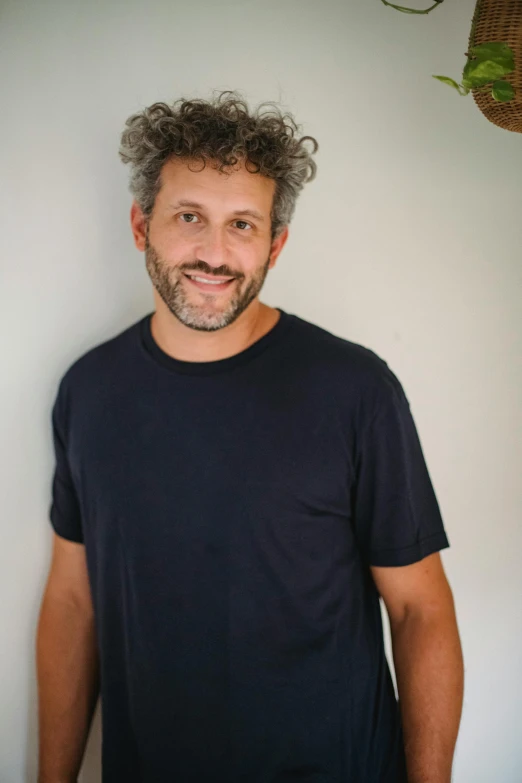 a man standing in front of a white wall, messy curly hair, dark grey haired man, riccardo federici, slightly smiling