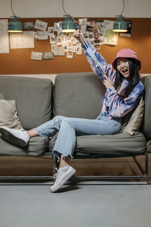a woman sitting on a couch with her arms in the air, inspired by Kim Jeong-hui, pexels contest winner, casual streetwear, gemma chan, wears a destroyed hat, blue jeans and grey sneakers