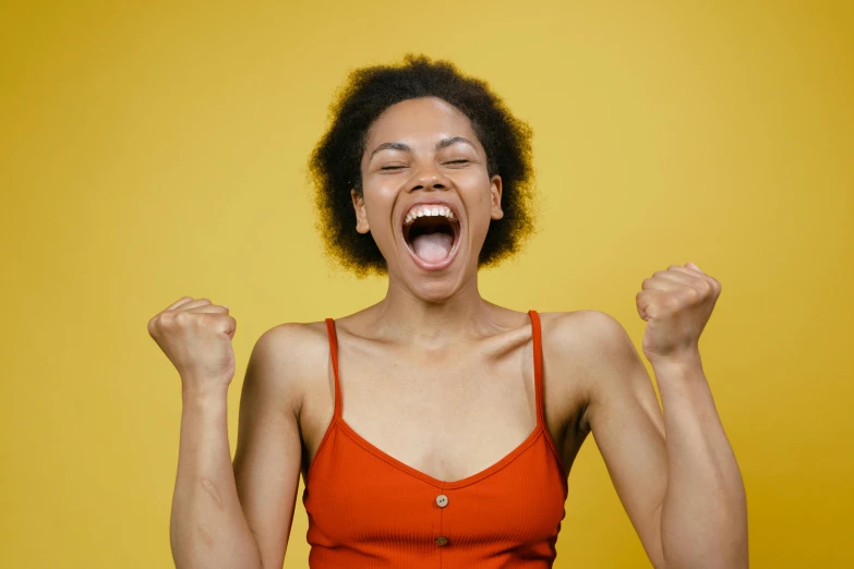 a woman in an orange top with her arms in the air, pexels contest winner, happening, clenching teeth, young black woman, yellow and red, victory