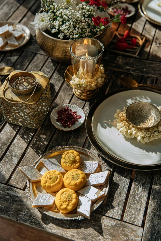 a wooden table topped with plates of food, inspired by Riad Beyrouti, hurufiyya, gold flaked flowers, sweets, outdoors setting, holiday season