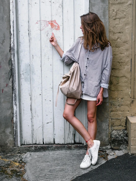 a woman standing in front of a white door, a picture, by Sara Saftleven, trending on unsplash, wear's beige shirt, wearing a baggy, grey and silver, wearing shorts