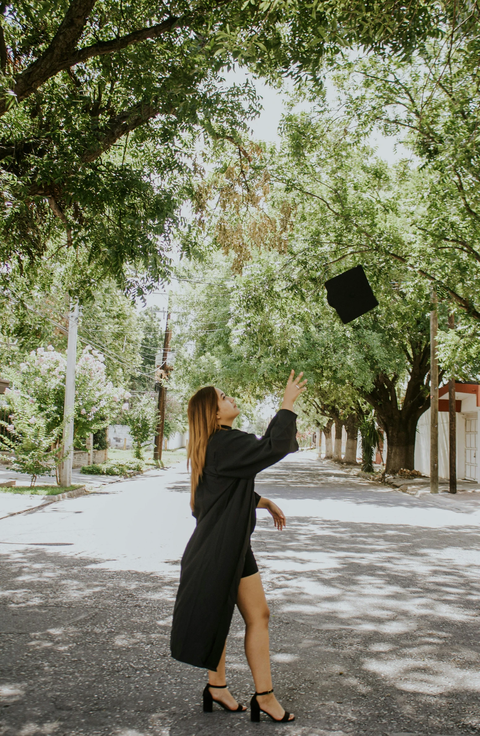a woman throwing a graduation hat in the air, a picture, pexels contest winner, tree in the background, walking to the right, black gown, street view