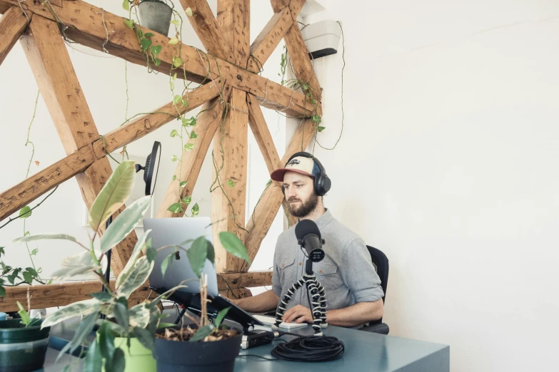 a man sitting at a desk in front of a computer, by Sebastian Vrancx, trending on unsplash, sitting in front of a microphone, in a white boho style studio, aussie baristas, covered in plants