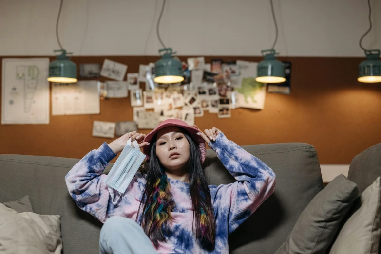 a woman sitting on a couch in a room, a portrait, inspired by helen huang, trending on pexels, hat and hoodie, tie-dye, young asian girl, outfit photo