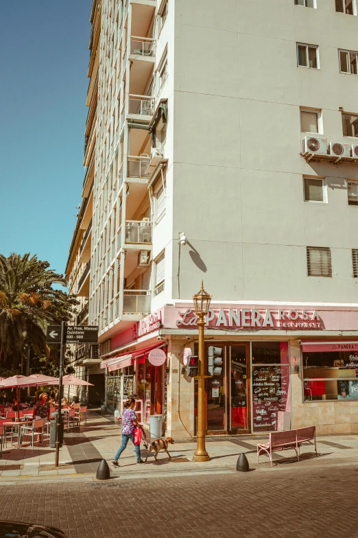 a couple of people walking down a street next to a tall building, marbella, bakery, buenos aires, sunfaded