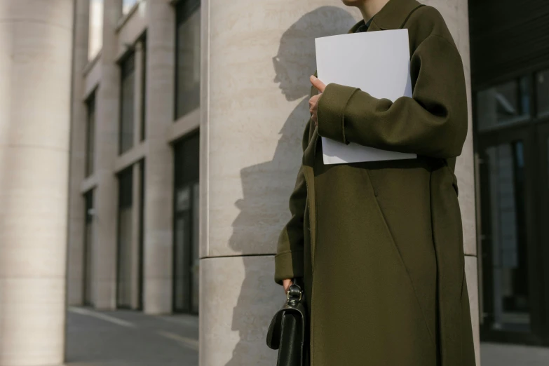 a woman standing in front of a building holding a folder, by Emma Andijewska, pexels contest winner, trench coat, olive green, a portrait of issey miyake, over-the-shoulder shot