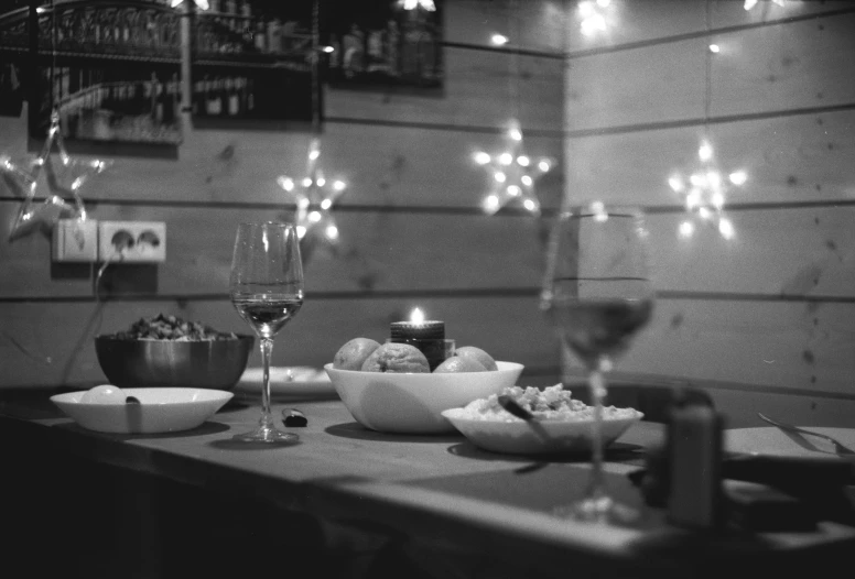 a table topped with bowls of food and glasses of wine, a black and white photo, by Caroline Mytinger, lights, 15081959 21121991 01012000 4k, holga 120n, festive atmosphere