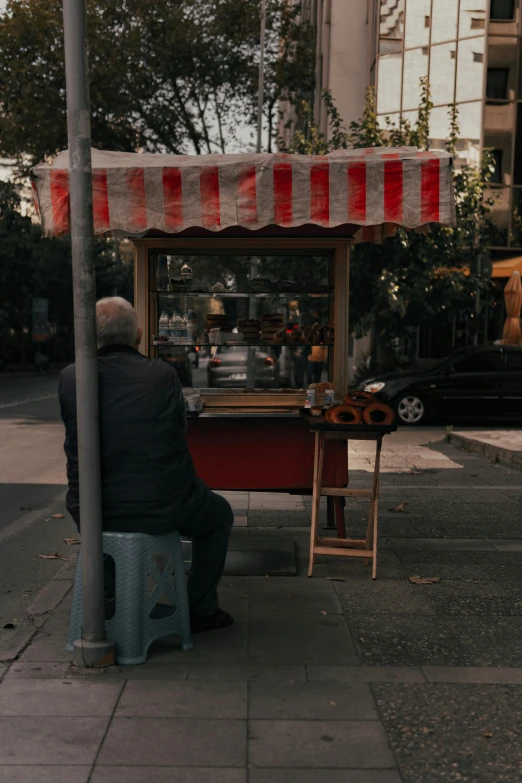 a man sitting on a stool on the side of a street, by Niko Henrichon, unsplash contest winner, food stall, popcorn machine, late morning, tel aviv street