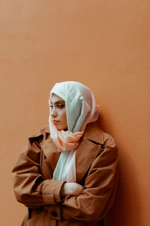 a woman leaning against a wall with her arms crossed, trending on pexels, hurufiyya, head scarf, brown jacket, pastel', lonely