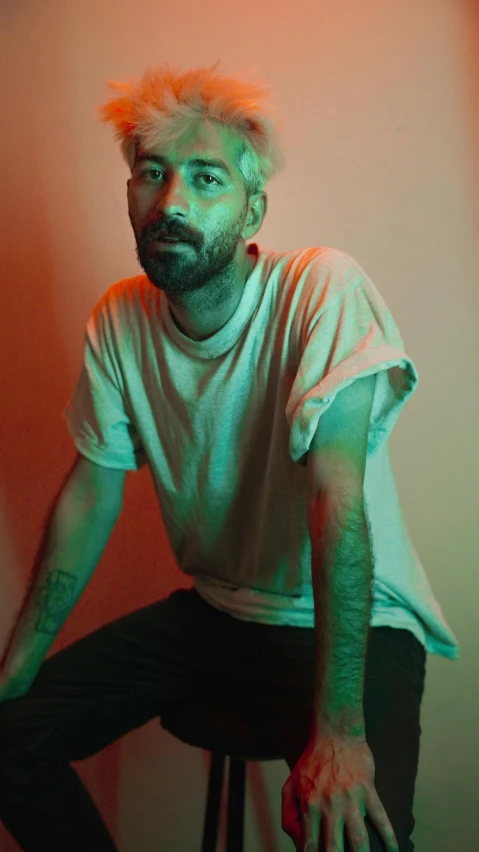 a man sitting on top of a stool, an album cover, inspired by Elsa Bleda, pexels, photorealism, light stubble with red shirt, light beard, james jean style, lgbtq