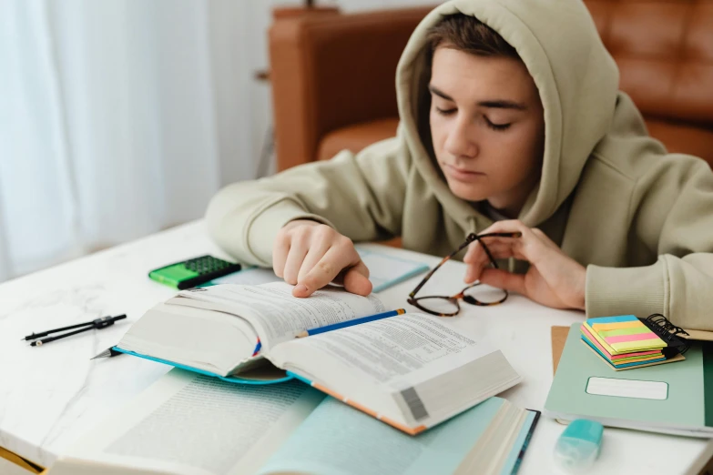 a person sitting at a table with a book and glasses, in a hoodie, educational supplies, teenage boy, trending on attestation