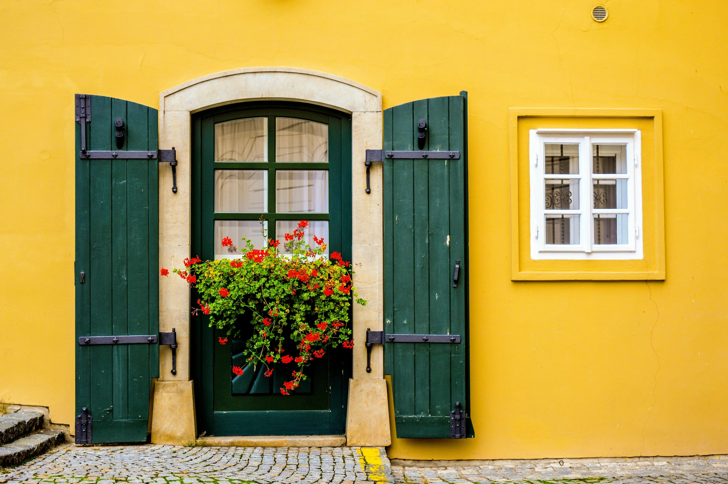 a yellow house with green shutters and red flowers, a photo, by Matthias Weischer, pexels contest winner, arts and crafts movement, green and gold, doorway, colors: yellow, small windows