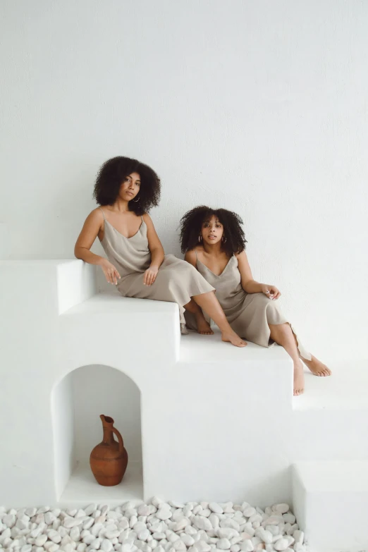 a couple of women sitting on top of a white wall, by Jessie Algie, trending on pexels, renaissance, brown skin like soil, organic dress, taupe, official store photo