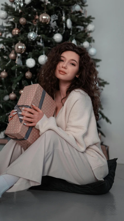 a woman sitting in front of a christmas tree holding a present, a colorized photo, by Emma Andijewska, pexels, renaissance, medium - long curly brown hair, doja cat, grey robe, wearing casual clothing