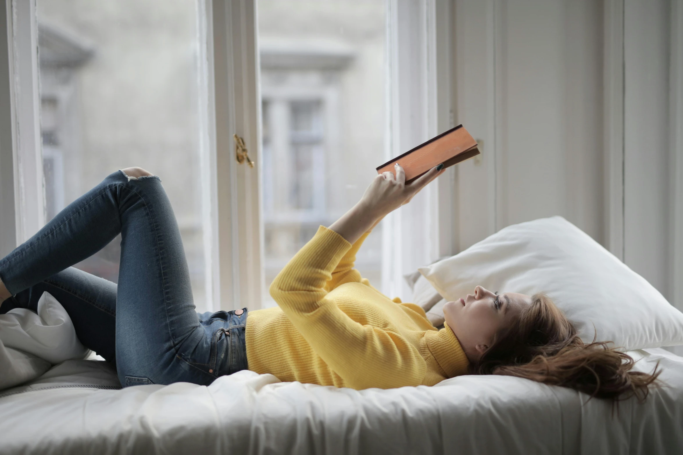 a woman laying on a bed reading a book, trending on pexels, with yellow cloths, next to window, casually dressed, super high resolution