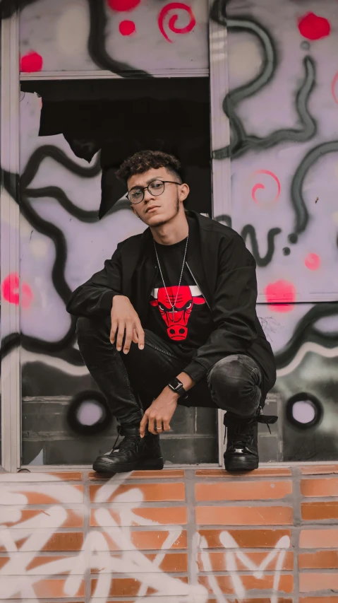 a man sitting on a ledge in front of a graffiti covered wall, an album cover, inspired by Ion Andreescu, pexels, red horns, casual black clothing, standing with a black background, an ox