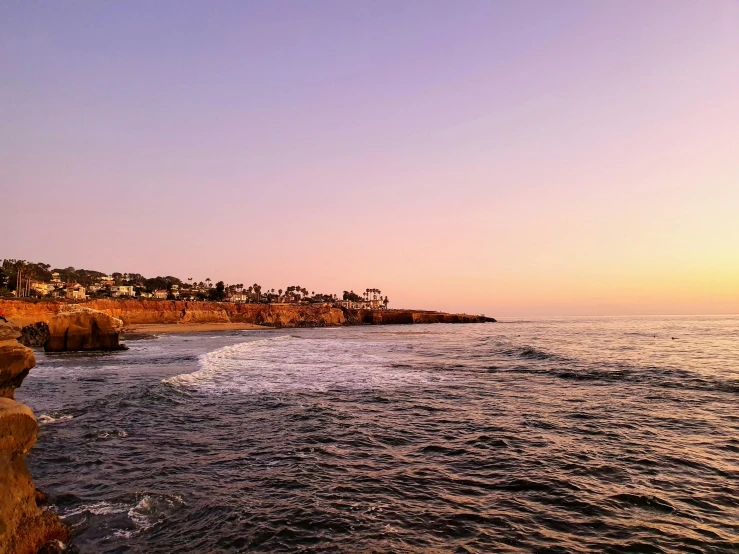 a man standing on top of a cliff next to the ocean, by Ryan Pancoast, pexels contest winner, vista of a city at sunset, pink hues, martin sandiego, panoramic