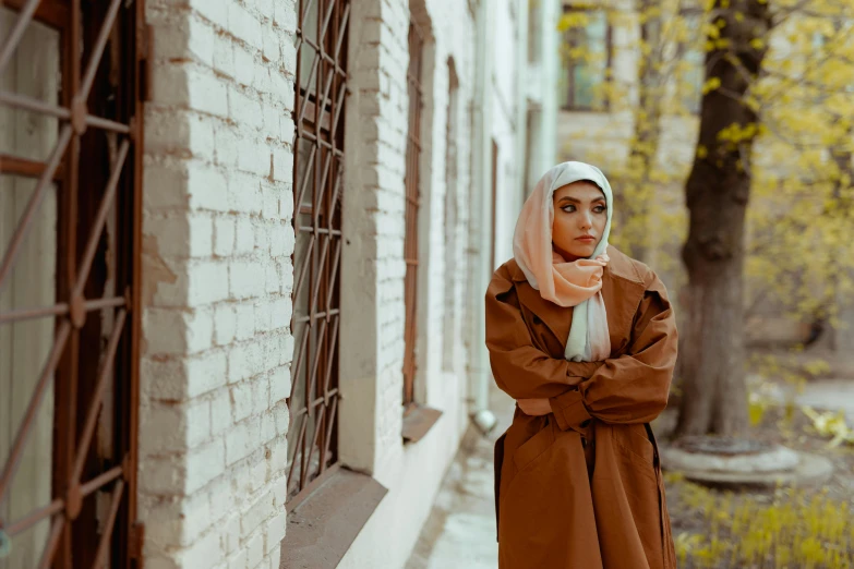 a woman standing in front of a brick building, pexels contest winner, hurufiyya, wearing a long coat, wearing a head scarf, brown jacket, concerned