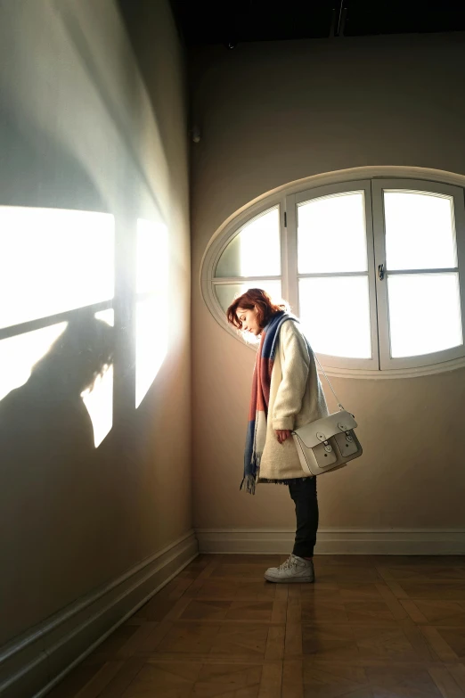 a woman standing in front of a window in a room, inspired by Nan Goldin, trending on pexels, light and space, studio ghibli sunlight, wearing a white winter coat, standing under a beam of light, looking sad