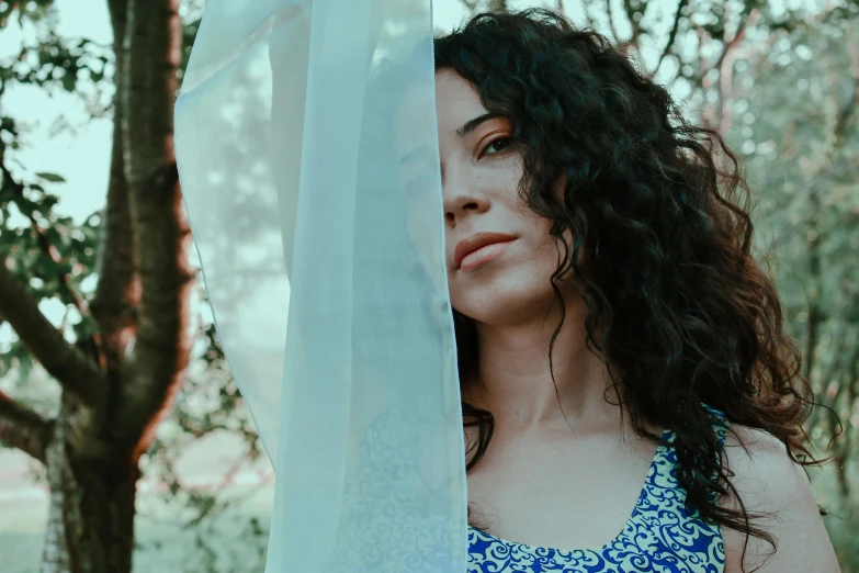 a woman wearing a blue dress standing in front of a tree, an album cover, inspired by Elsa Bleda, pexels contest winner, curly haired, pensive expression, young middle eastern woman, looking outside