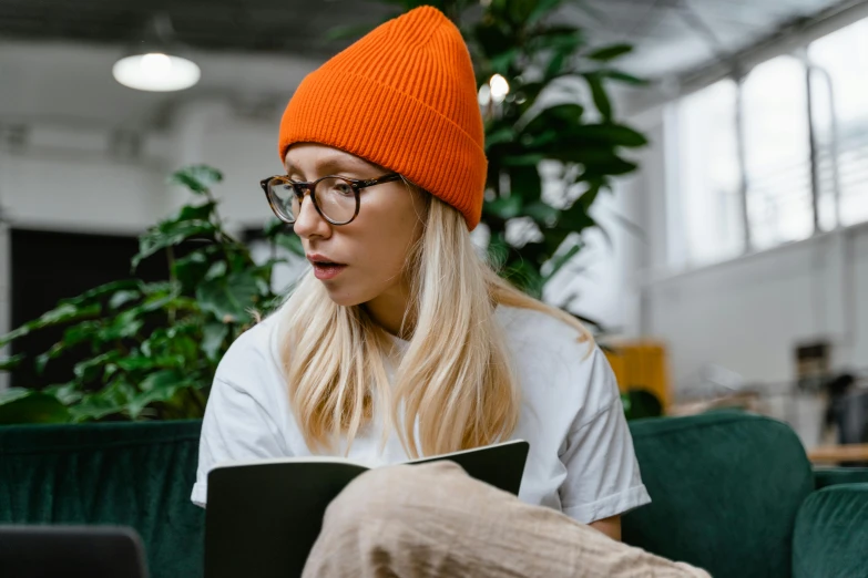 a woman sitting on a couch using a laptop, a cartoon, trending on pexels, beanie, reading engineering book, orange hoodie, academic clothing