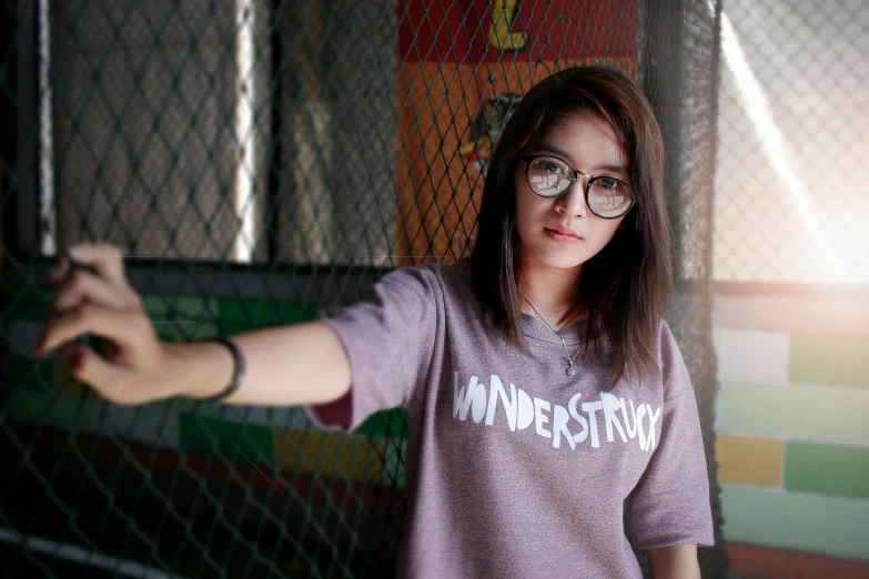 a woman with glasses standing in front of a fence, inspired by Rudy Siswanto, trending on unsplash, graffiti, in tshirt, wonderous, studio photo, with index finger