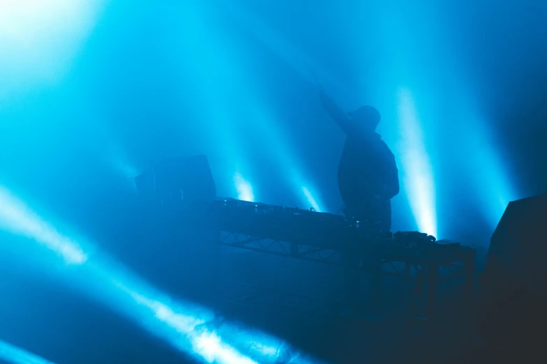 a man that is standing in front of a keyboard, unsplash, light and space, scene from a rave, blue mist, on stage, poorly lit