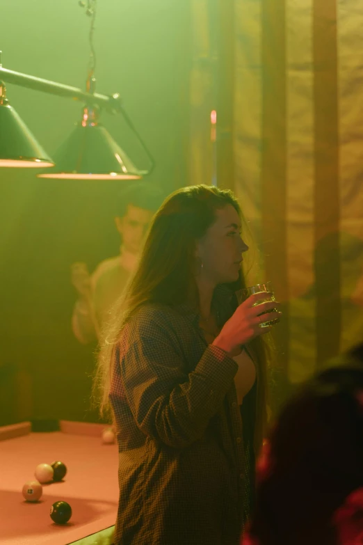 a woman standing in front of a pool table, a portrait, inspired by Nan Goldin, unsplash, drinking whiskey, yellow lighting from right, many partygoers, ( ( theatrical ) )