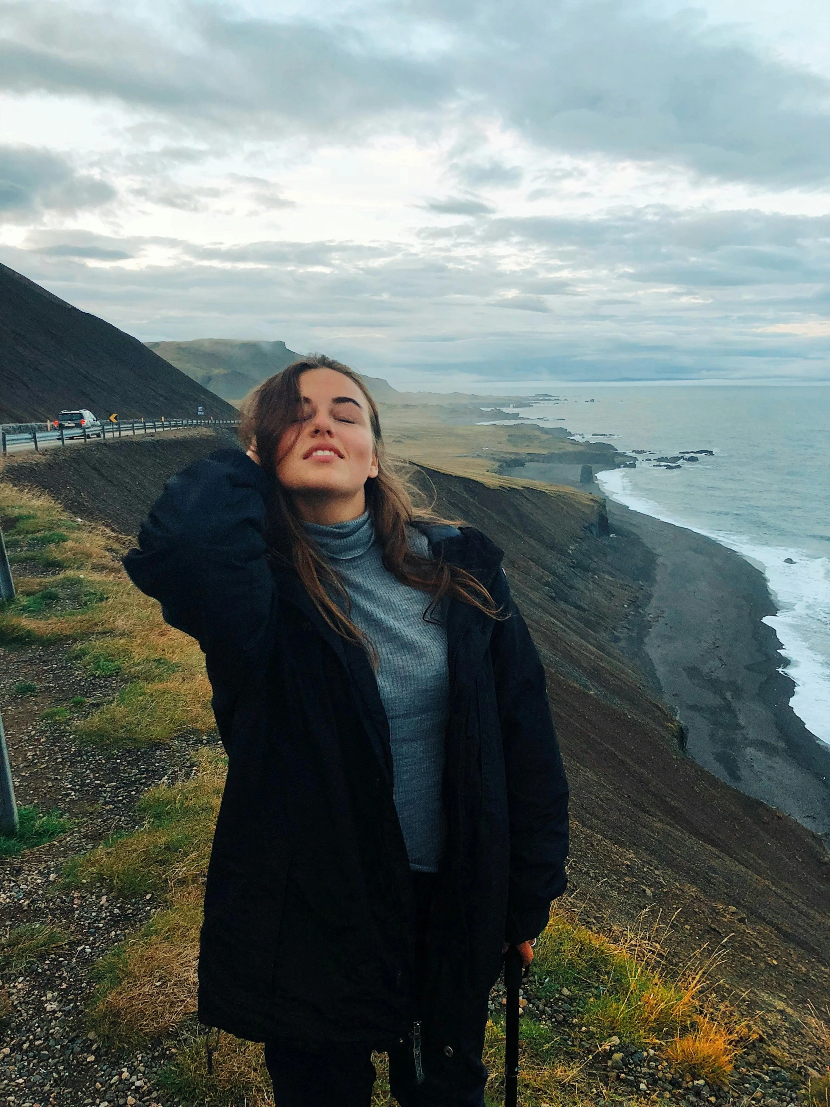 a woman standing on top of a cliff next to the ocean, wearing a turtleneck and jacket, ☁🌪🌙👩🏾, russian girlfriend, wavy hair spread out