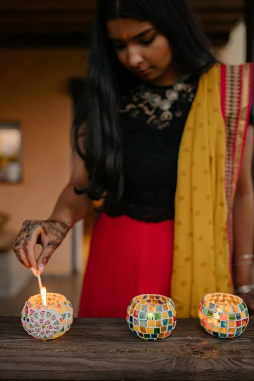 a woman lighting a candle on a table, by Pacita Abad, trending on unsplash, elegantly dressed, indian, glowing accents, 15081959 21121991 01012000 4k