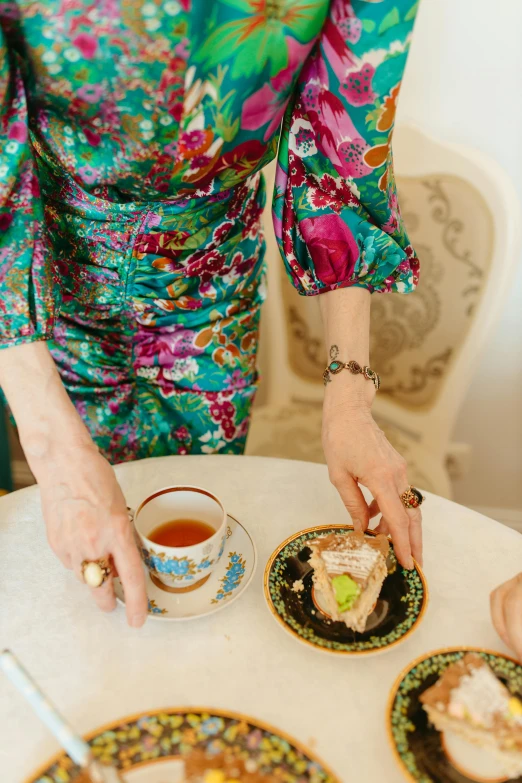 a woman sitting at a table with plates of food, by Julia Pishtar, trending on pexels, rococo, patterned clothing, tea cup, high angle close up shot, soft silk dress