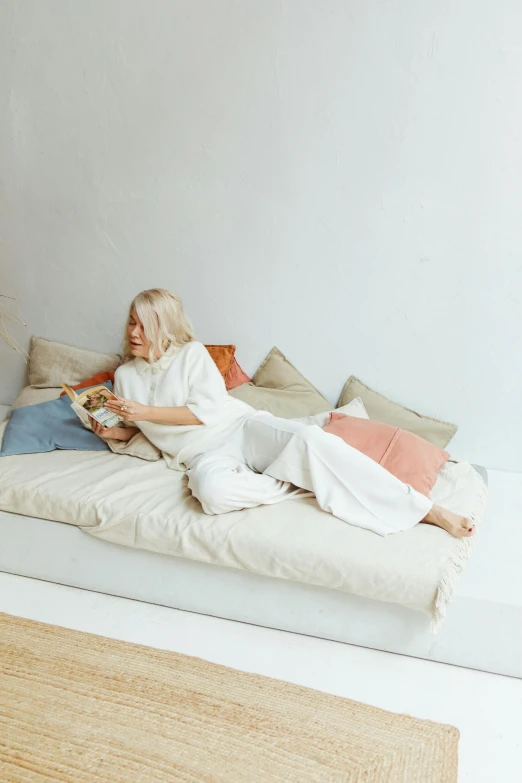 a woman sitting on a bed reading a book, inspired by Louisa Matthíasdóttir, in a white boho style studio, sitting on couch, a blond, uploaded