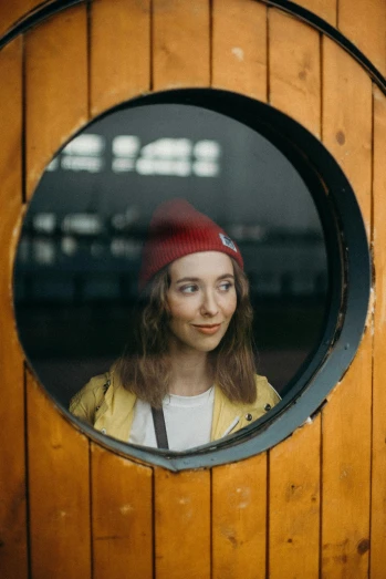 a woman is looking out of a round window, inspired by Wes Anderson, pexels contest winner, wearing a beanie, good looking face, anna nikonova, wearing a scarlet hoodie