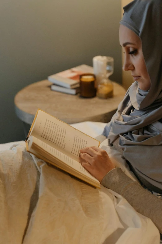 a woman in a hijab is reading a book, someone sits in bed, wearing a grey robe, books on side table, profile image