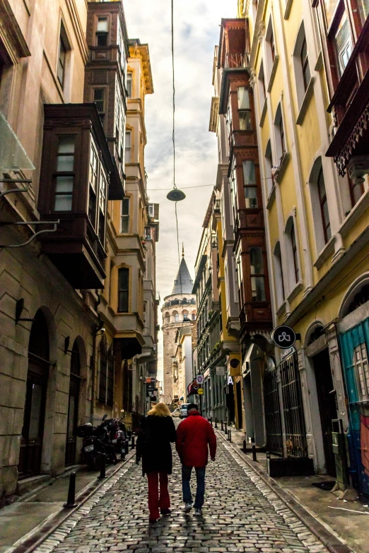 two people walking down a cobblestone street, by Cafer Bater, pexels contest winner, art nouveau, ottoman sultan, tall towers, view from bottom, wires hanging above street