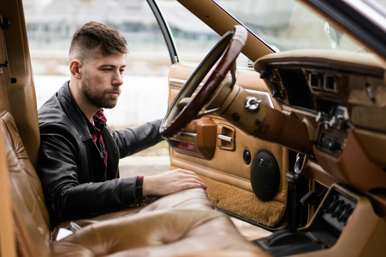 a man sitting in the driver's seat of a car, an album cover, by Julia Pishtar, auto-destructive art, bmw m1 lincoln continental, dusty rown bomber leather jacket, thumbnail, high end interior