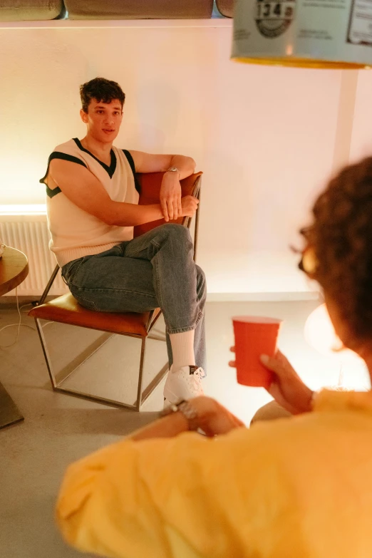 a man sitting in a chair in a room, teenager hangout spot, queer woman, holding a drink, looking at each other mindlessly