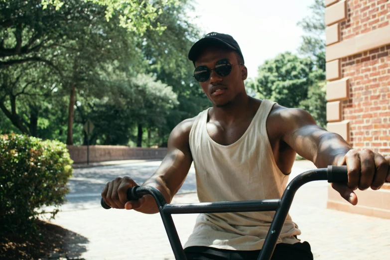 a man riding a bike next to a brick building, wearing a tank top and shorts, in style of tyler mitchell, wearing sunglasses and a hat, parks and gardens