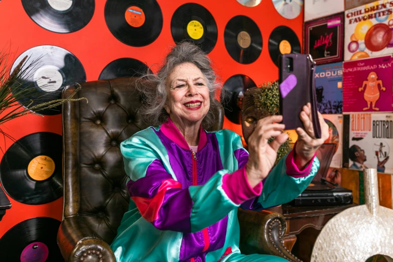 a woman sitting in a chair holding a cell phone, an album cover, maximalism, grandma, taking a selfie, wearing silver silk robe, bright trouser suit for a rave