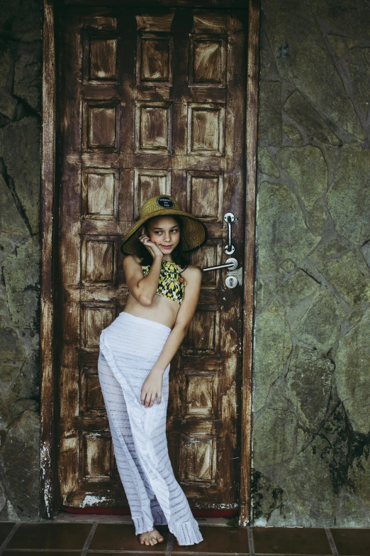 a woman standing in front of a wooden door, by Lucia Peka, unsplash, wearing seashell attire, caracter with brown hat, white sarong, cavewoman