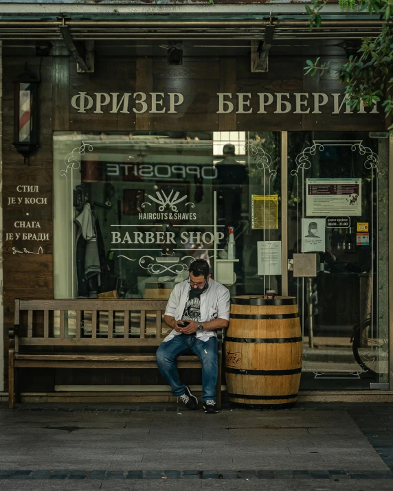 a man sitting on a bench in front of a barber shop, by andrei riabovitchev, pexels contest winner, barrel chested, bookshops, 000 — википедия, storefront