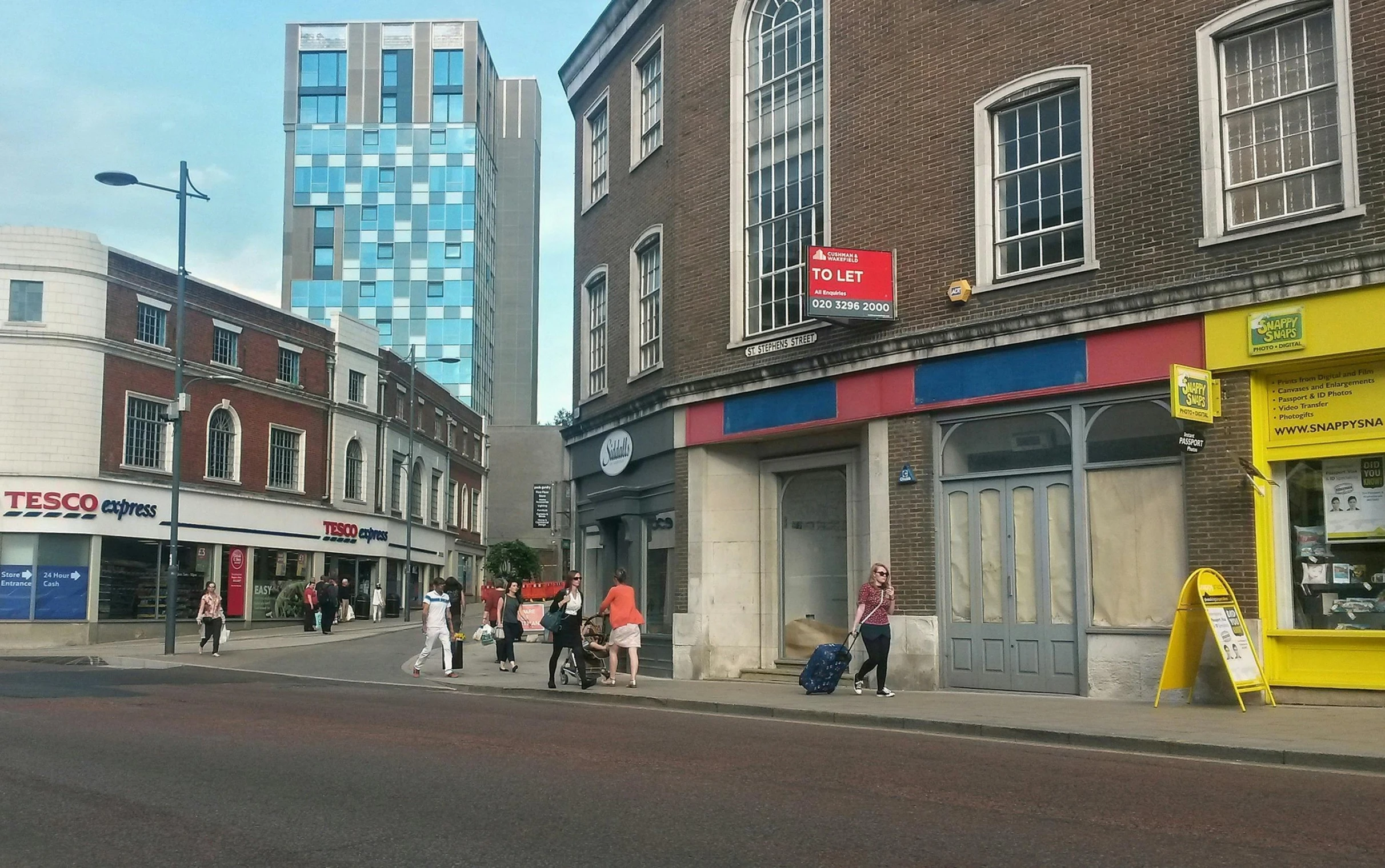 a group of people walking down a street next to tall buildings, an album cover, unsplash, hyperrealism, coventry city centre, fresh bakeries in the background, indigo and venetian red, photo taken with an iphone