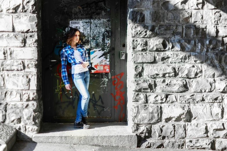 a woman standing in the doorway of a building, an album cover, inspired by Silvestro Lega, pexels contest winner, graffiti, blue jeans, wearing plaid shirt, promotional image, rock star