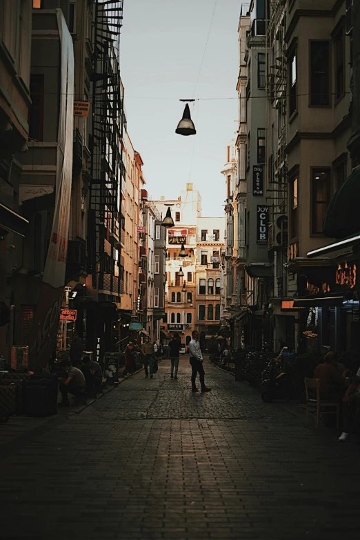 a couple of people walking down a street next to tall buildings, a photo, renaissance, turkey, warm street lights store front, square, unsplash 4k