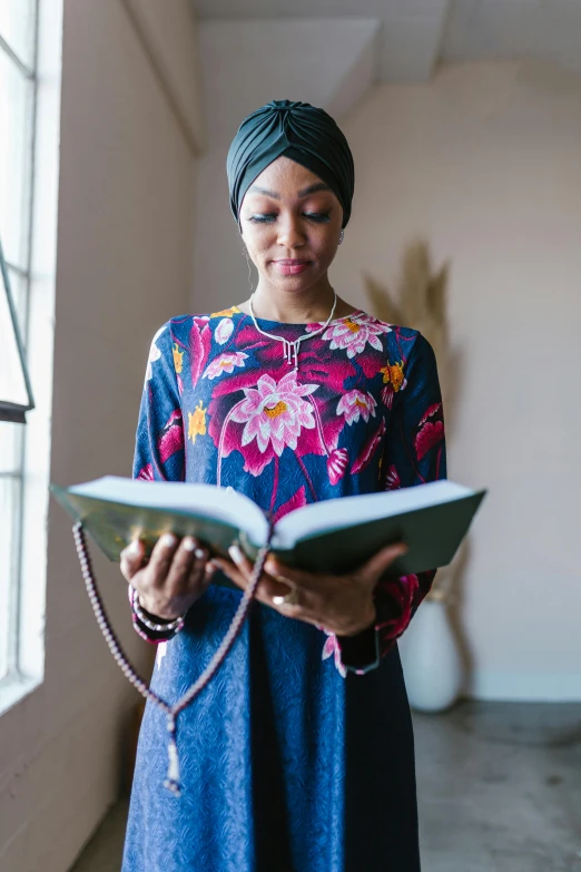 a woman standing in front of a window holding a book, inspired by Maryam Hashemi, pexels contest winner, hurufiyya, wearing a turban, african american woman, wearing cross on robe, reading