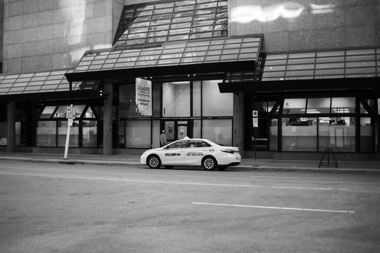 a white car parked in front of a building, a black and white photo, by Maurycy Gottlieb, pexels contest winner, vancouver school, security agent, exiting store, taxi, yeg