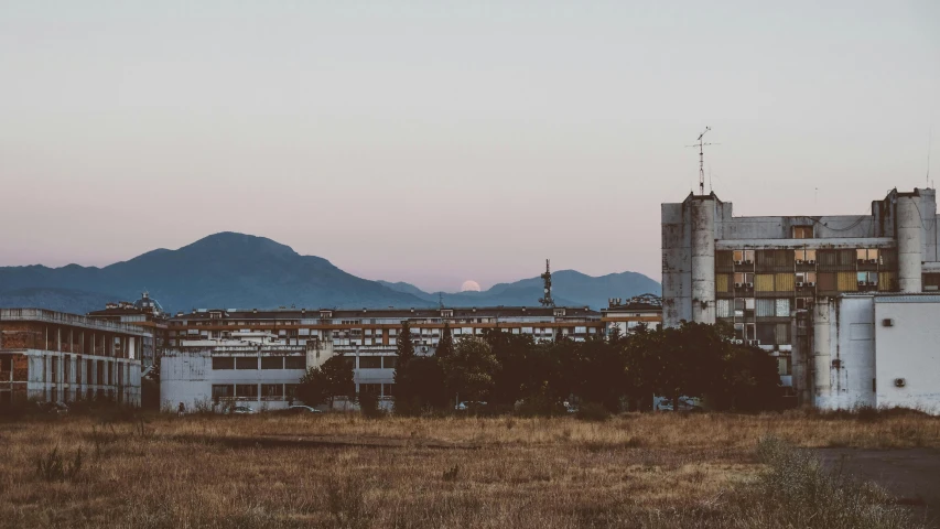 a field with buildings and mountains in the background, unsplash, brutalism, big moon in the background, industrial surrounding, southern european scenery, sunfaded