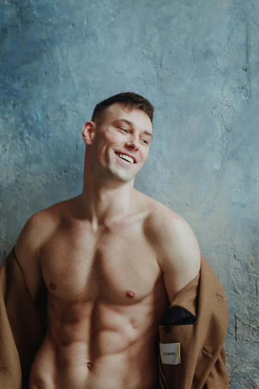 a shirtless man standing in front of a blue wall, an album cover, inspired by Ryan Pancoast, trending on pexels, smiling fashion model, he is wearing a trenchcoat, bare bodybuilder shoulders. kohl, androgynous person